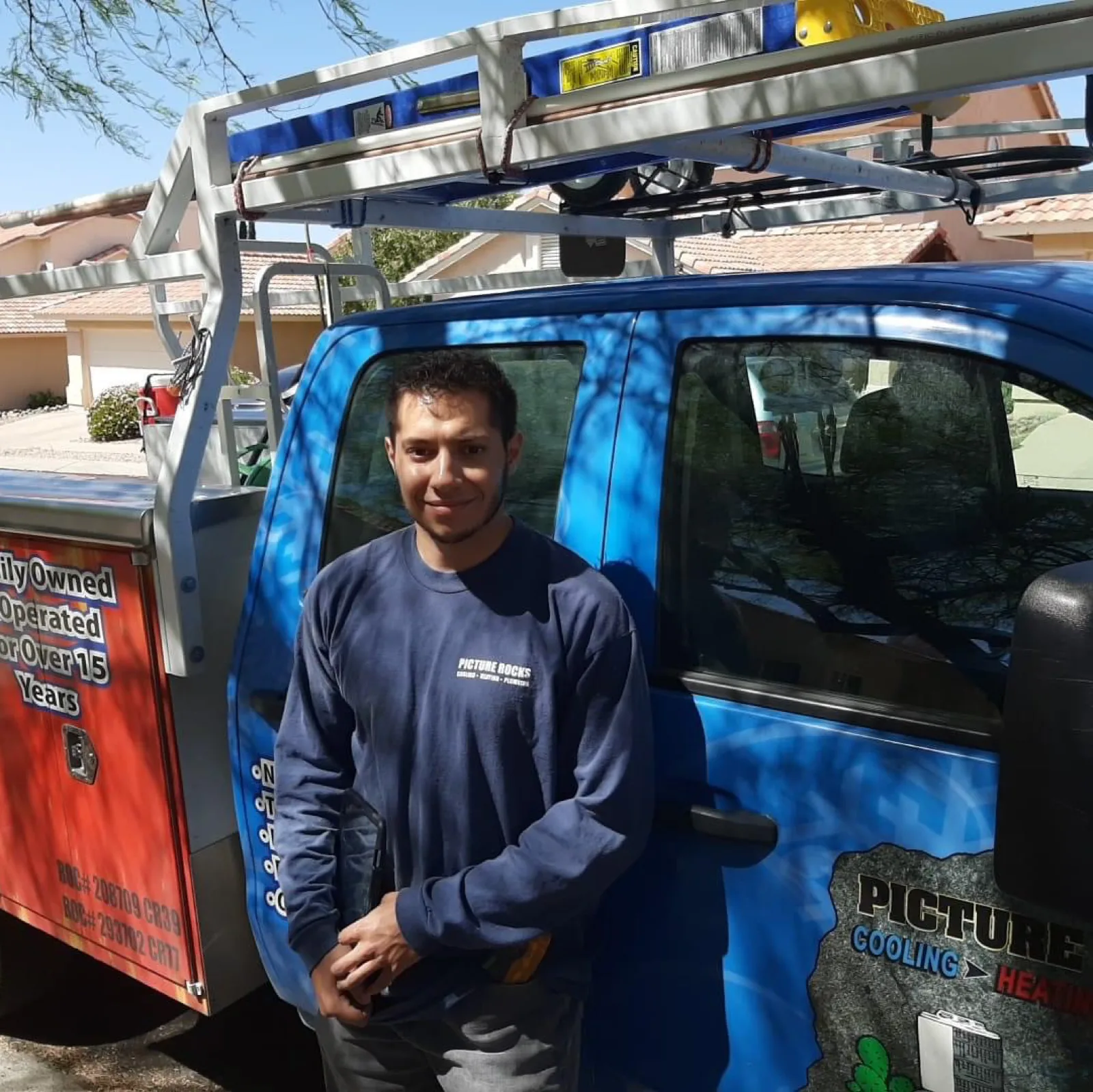 Picture Rocks Cooling technician standing beside a blue-wrapped service truck with a ladder rack.